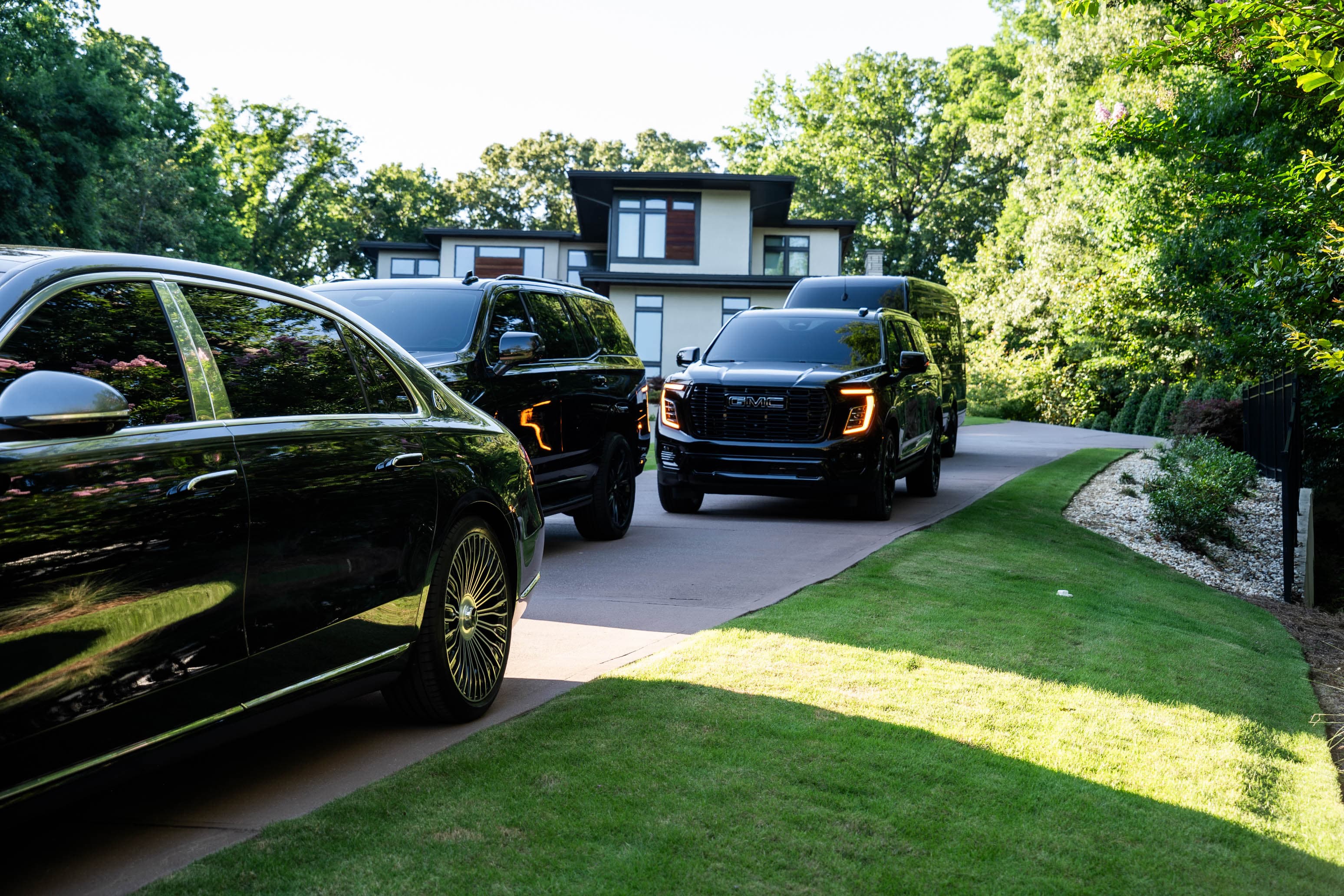 Black luxury vehicles parked on a driveway leading to a modern house with lush greenery.