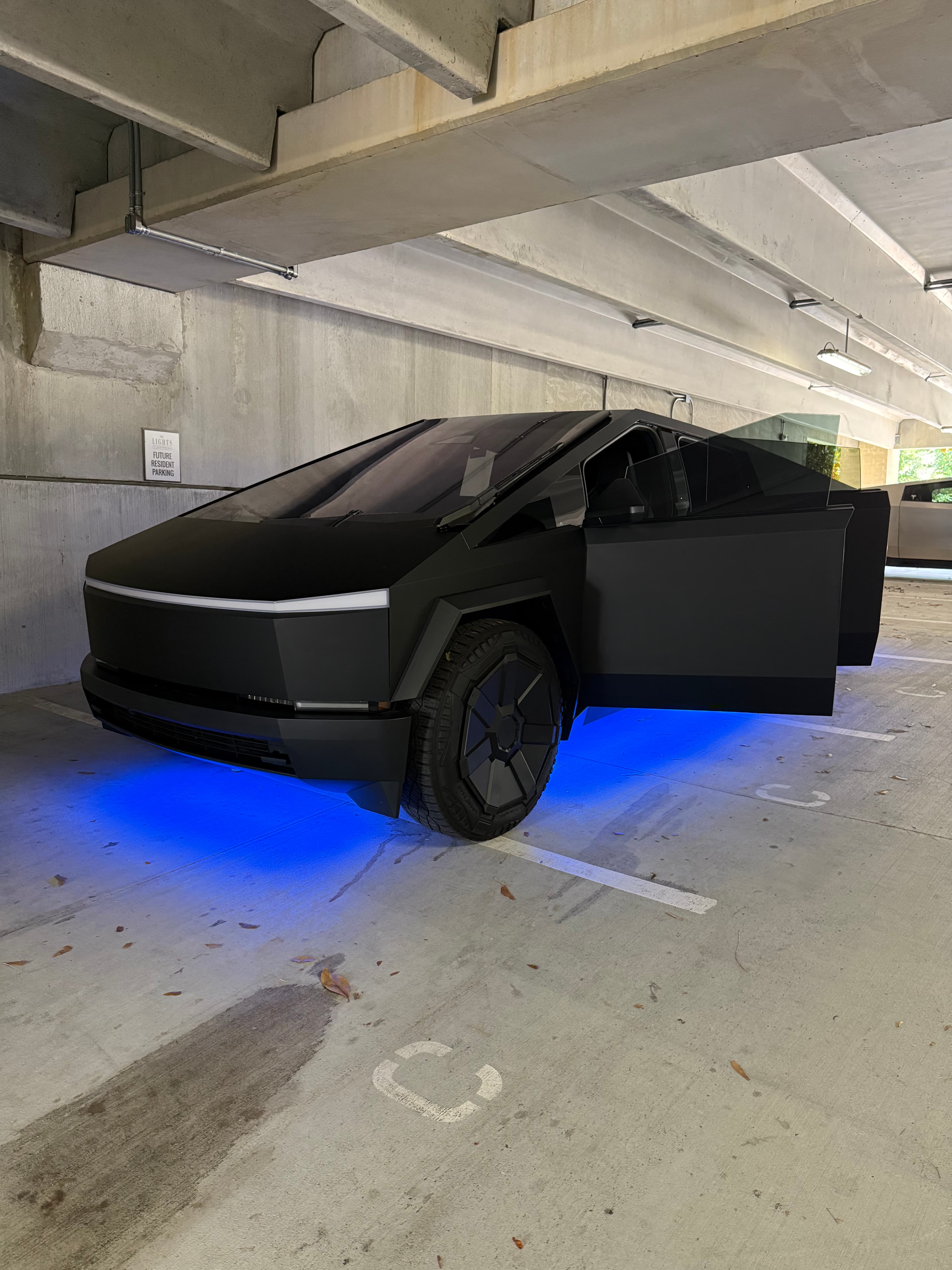 Matte black Tesla Cybertruck with blue underglow lighting and open door in a concrete garage.