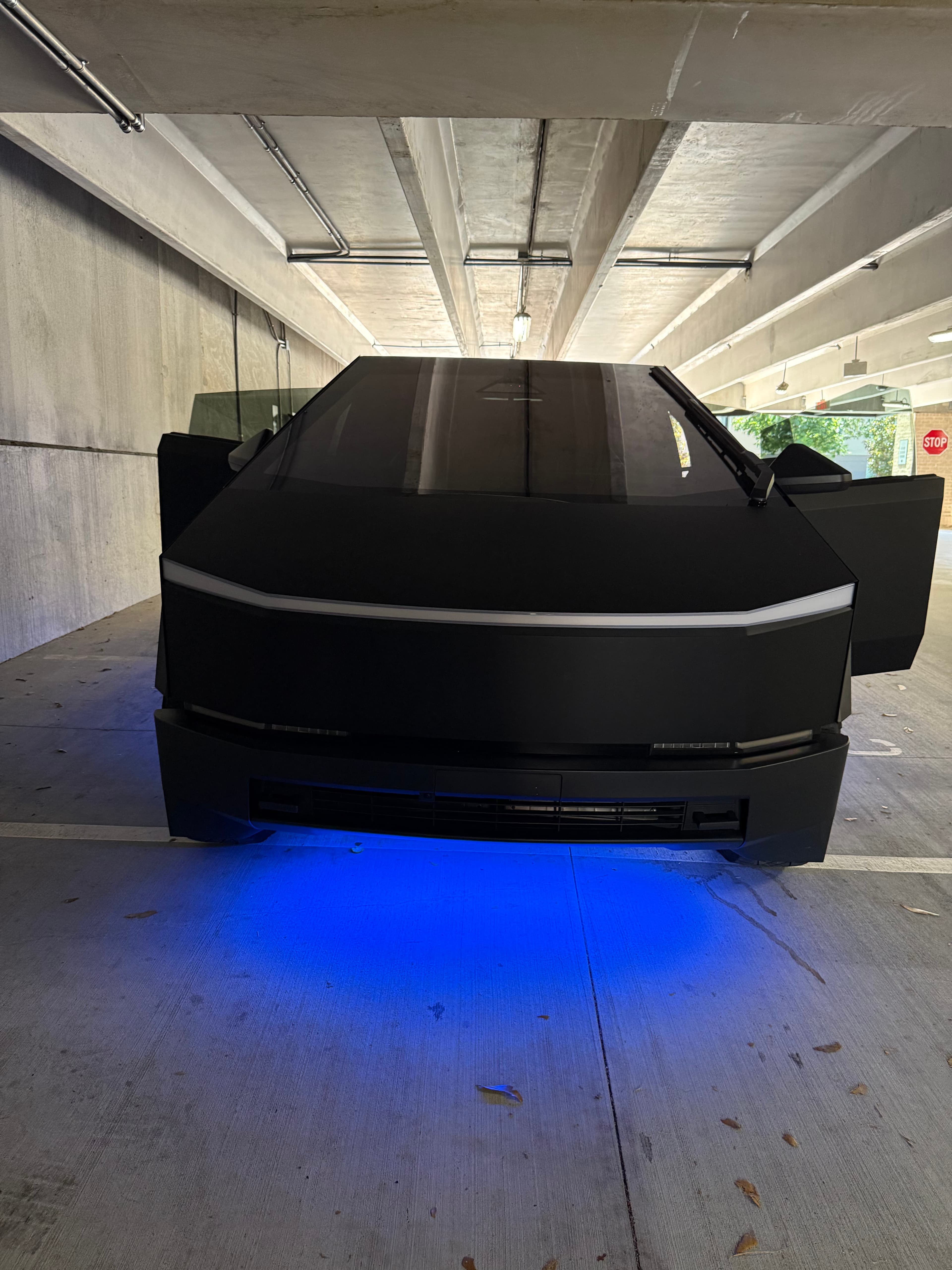 Matte black Cybertruck with open doors and vibrant blue underglow lighting in a parking garage.